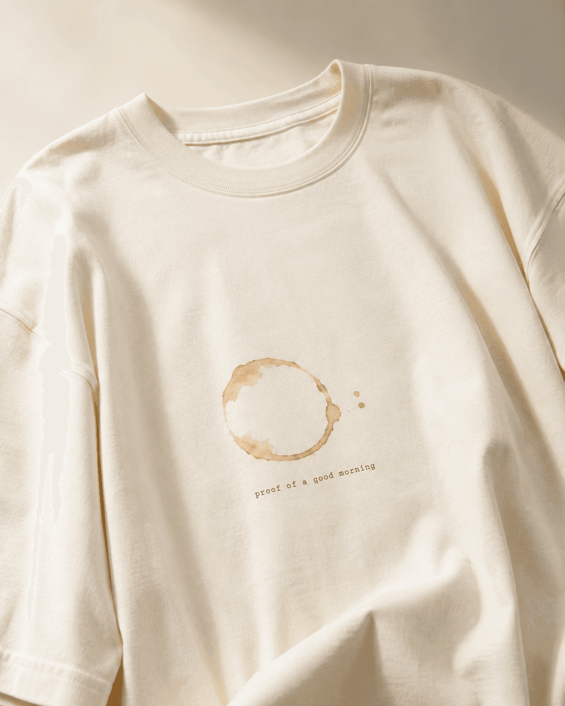 Close-up of the folded cream t-shirt's collar and chest, showing the faded coffee ring stain print and the caption 'proof of a good morning'.